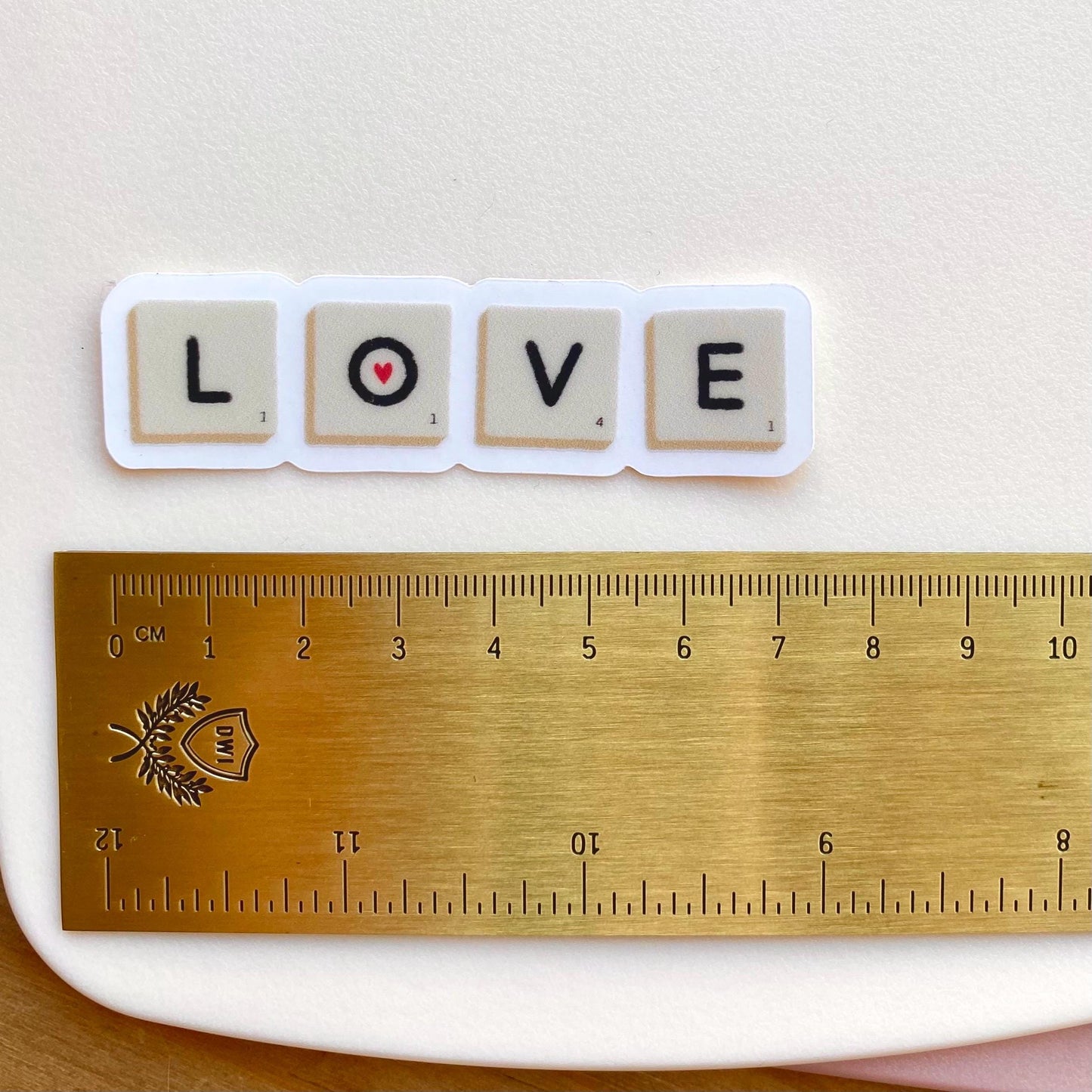 A sticker with Scrabble tiles spelling 'LOVE' on a white surface with a gold ruler for scale.