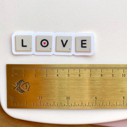 A sticker with Scrabble tiles spelling 'LOVE' on a white surface with a gold ruler for scale.
