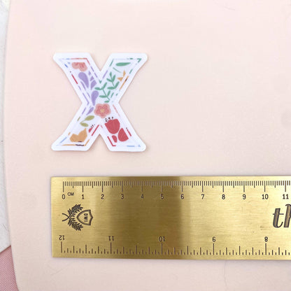 Floral letter 'X' sticker next to a gold ruler on a pink background