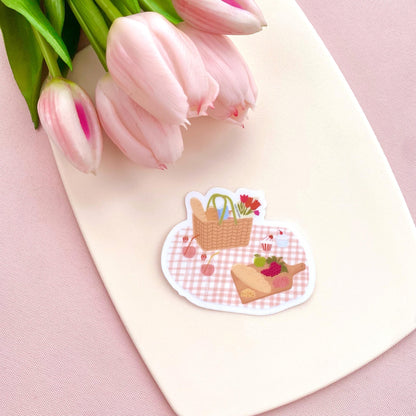 Sticker of a picnic scene with a basket and food on a white card with pink tulips.