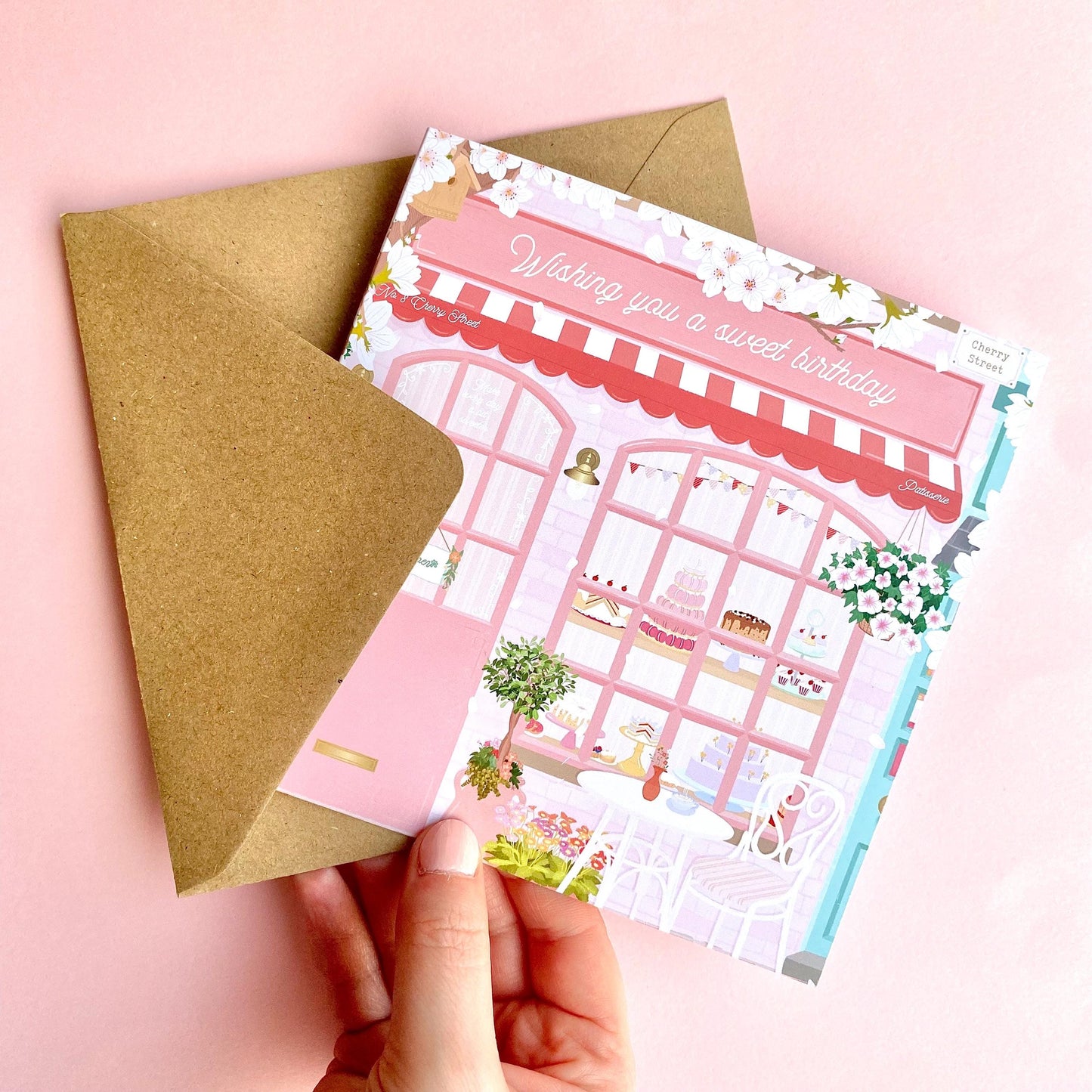 Hand holding a pink bakery birthday card with a brown kraft envelope on a pink background