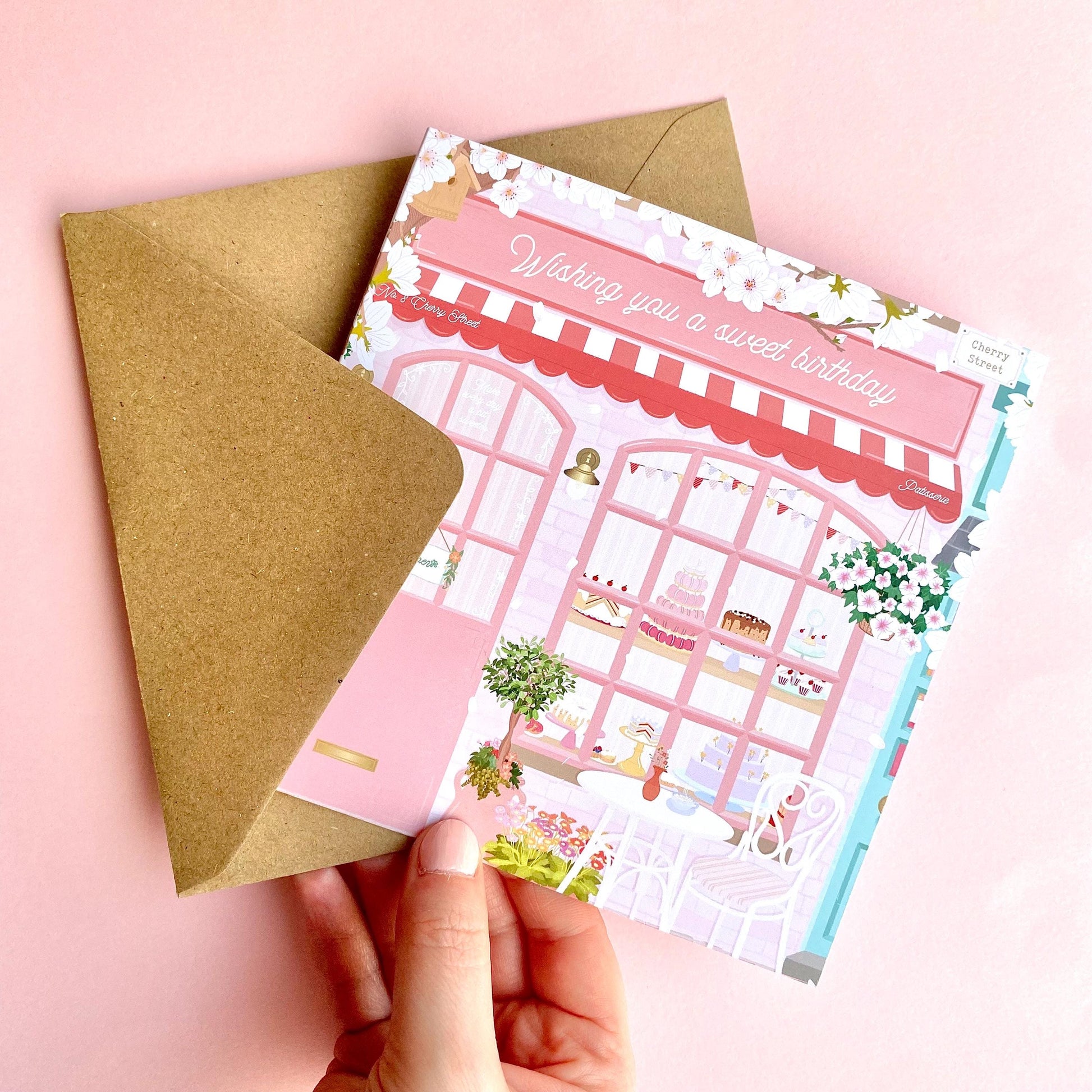 Hand holding a pink bakery birthday card with a brown kraft envelope on a pink background