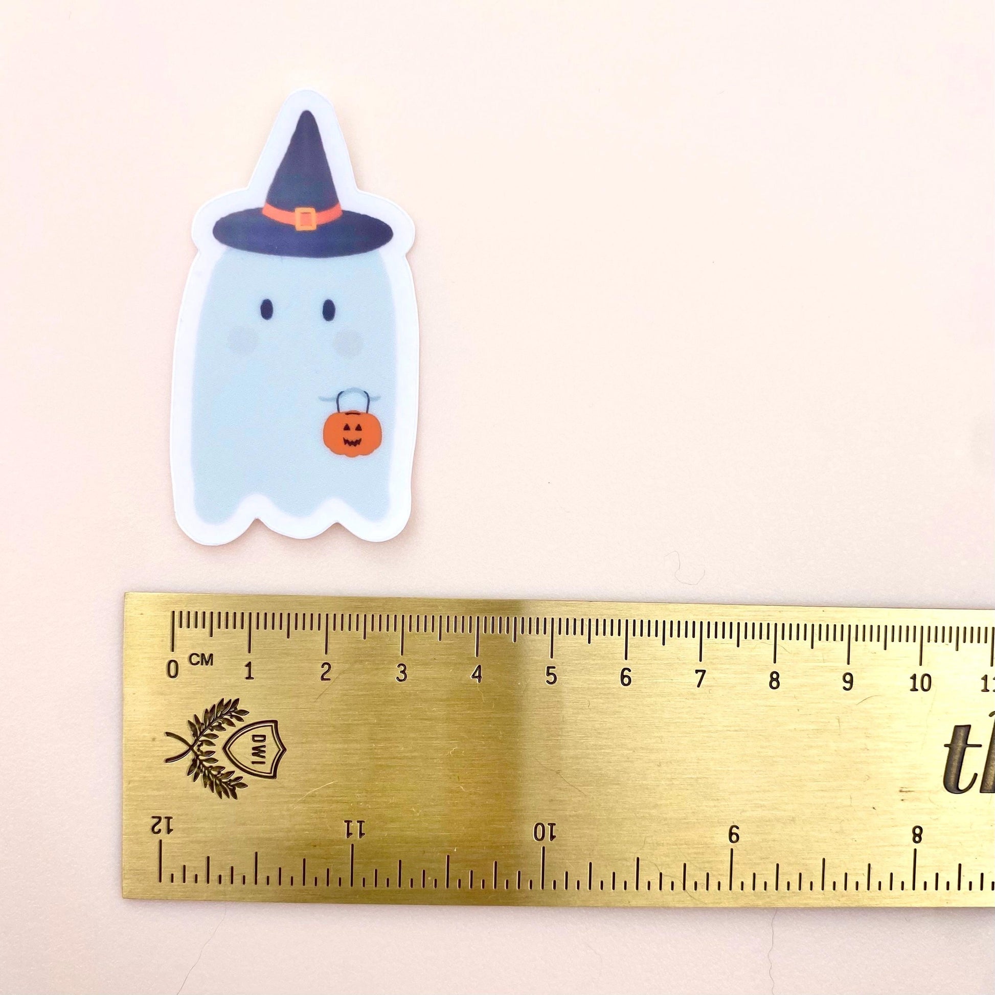Halloween-themed ghost sticker with a witch hat and pumpkin next to a ruler for scale on a light pink background.