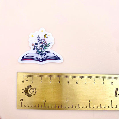 Decorative sticker of an open book with floral designs and a moon and stars on a white background with a gold ruler for scale