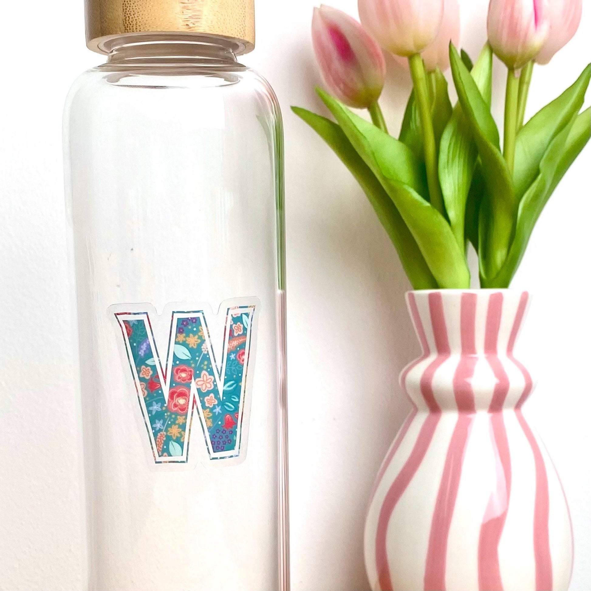 Clear water bottle with floral letter 'W' sticker next to a striped vase with tulips on a white background