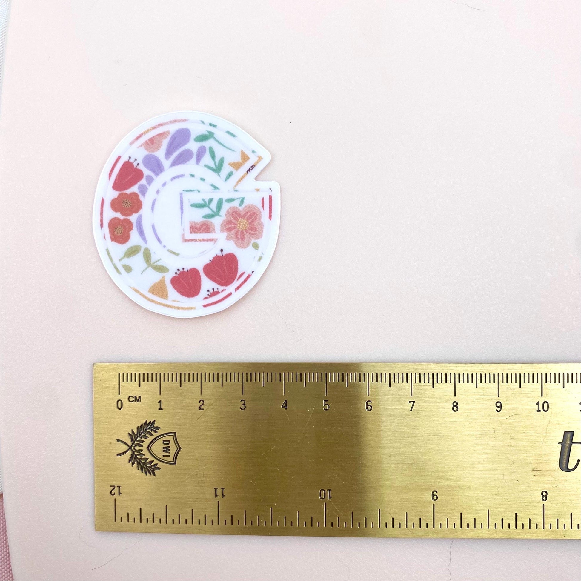 Decorative letter 'G' sticker with floral design on a pink background, next to a gold ruler for scale.