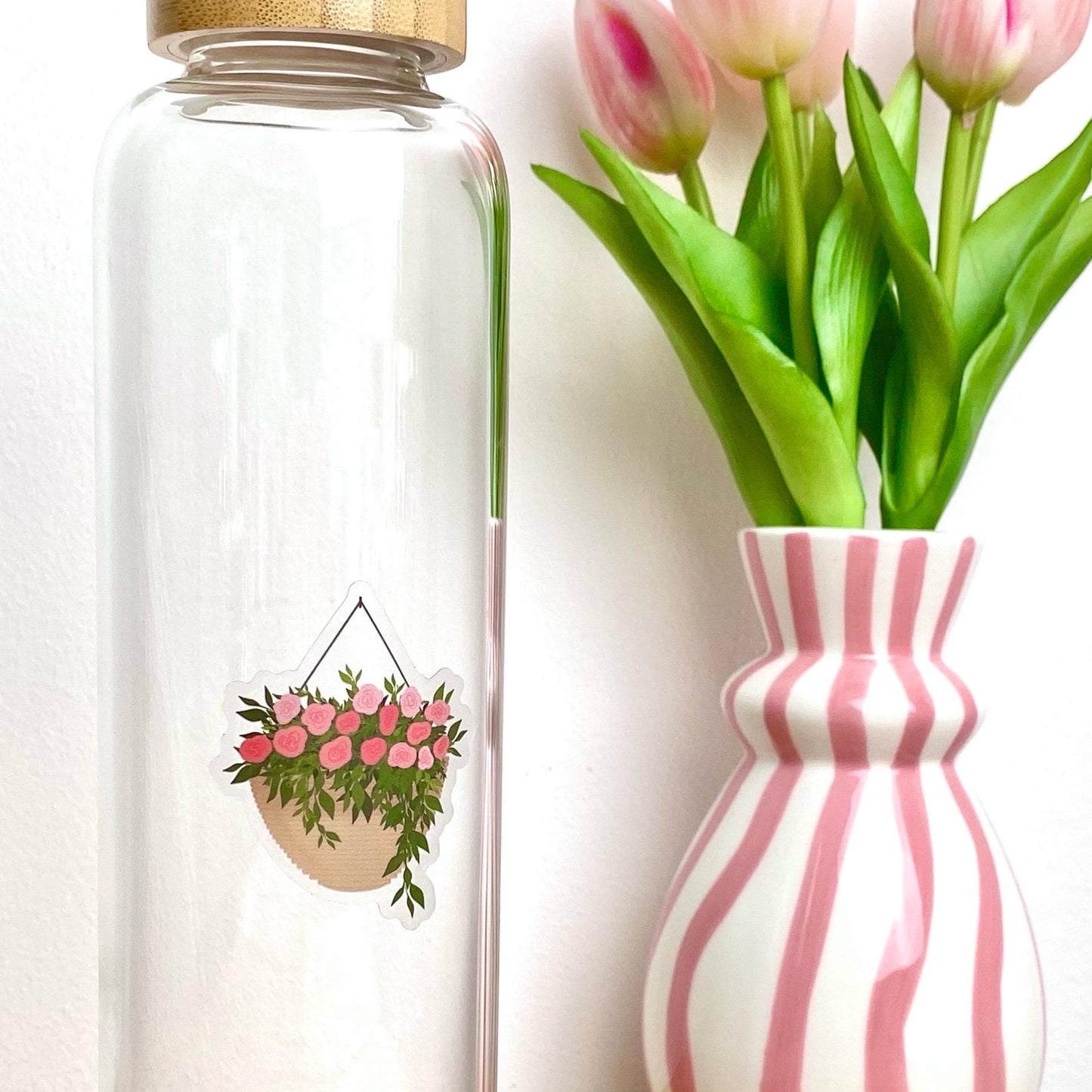 A sticker with a hanging planter with pink flowers against a glass bottle besides a pink striped vase of tulips.