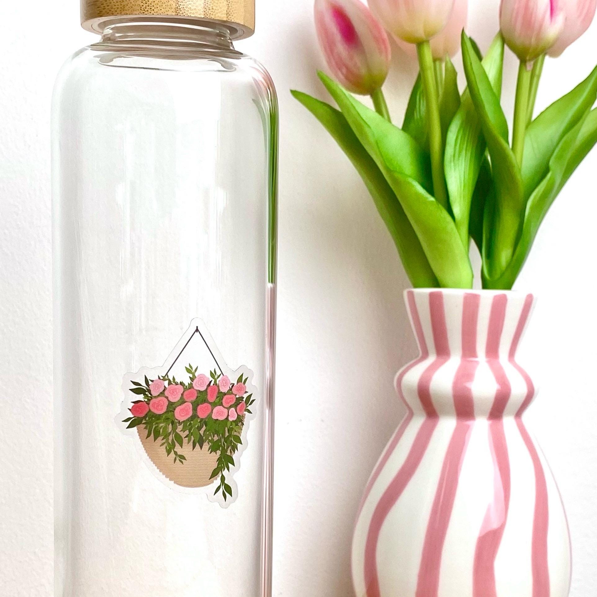 A sticker with a hanging planter with pink flowers against a glass bottle besides a pink striped vase of tulips.