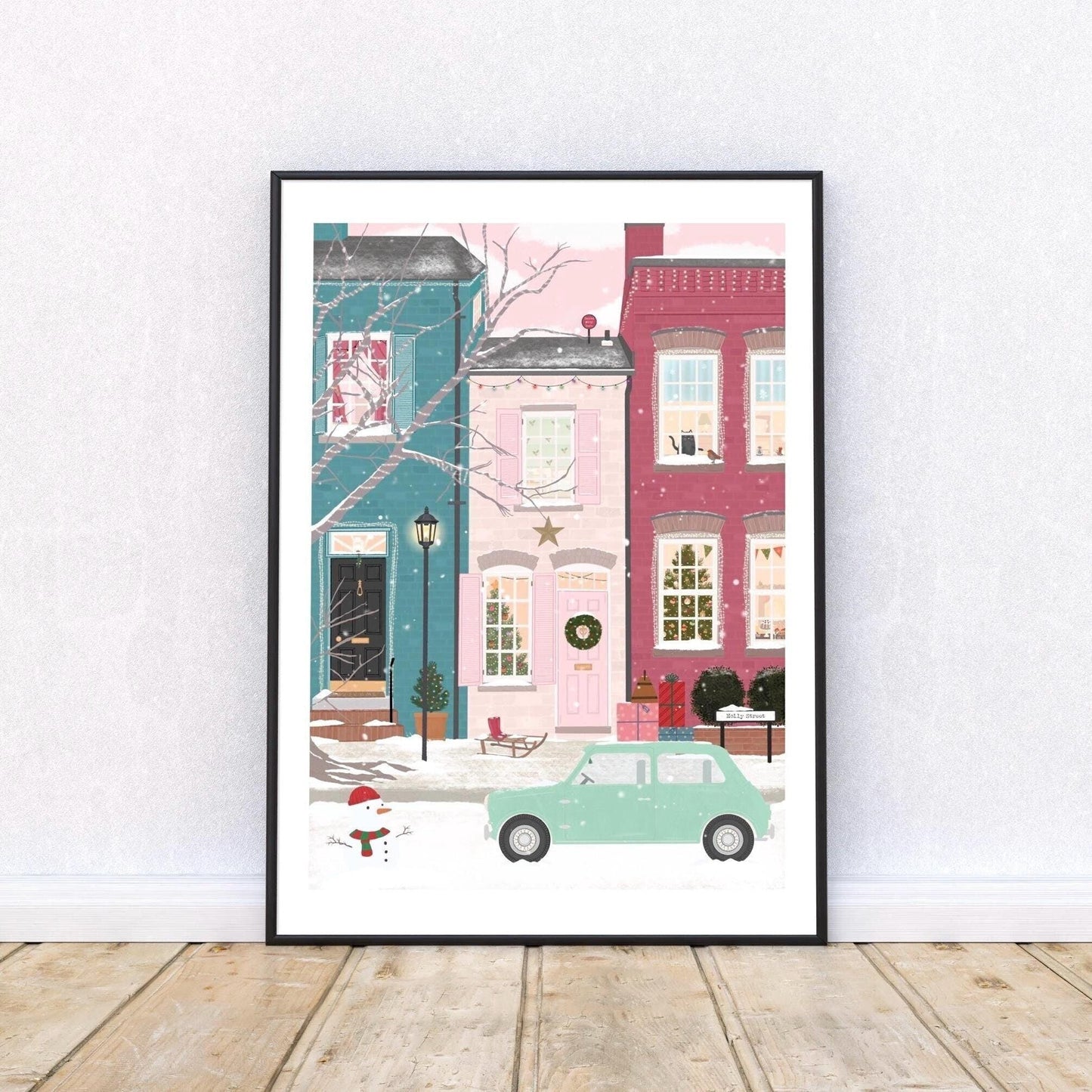 Framed artwork of a snowy street scene with colorful houses and a green car on a white wall.