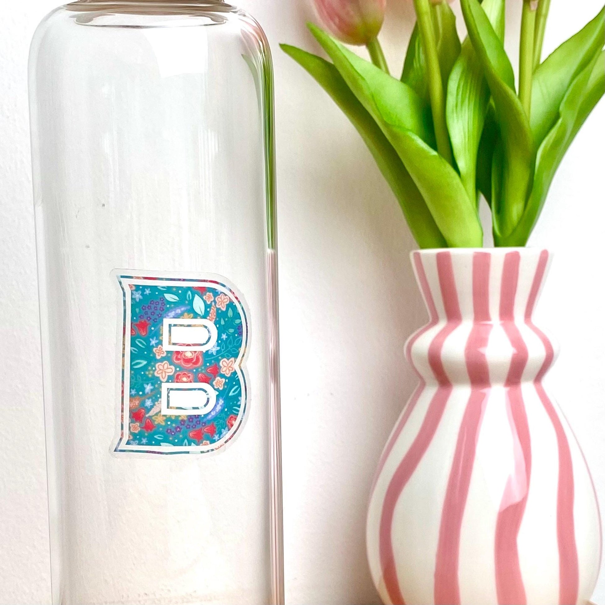 Clear sticker with a teal floral decorated letter 'B' against a glass bottle besides a pink striped vase of tulips.