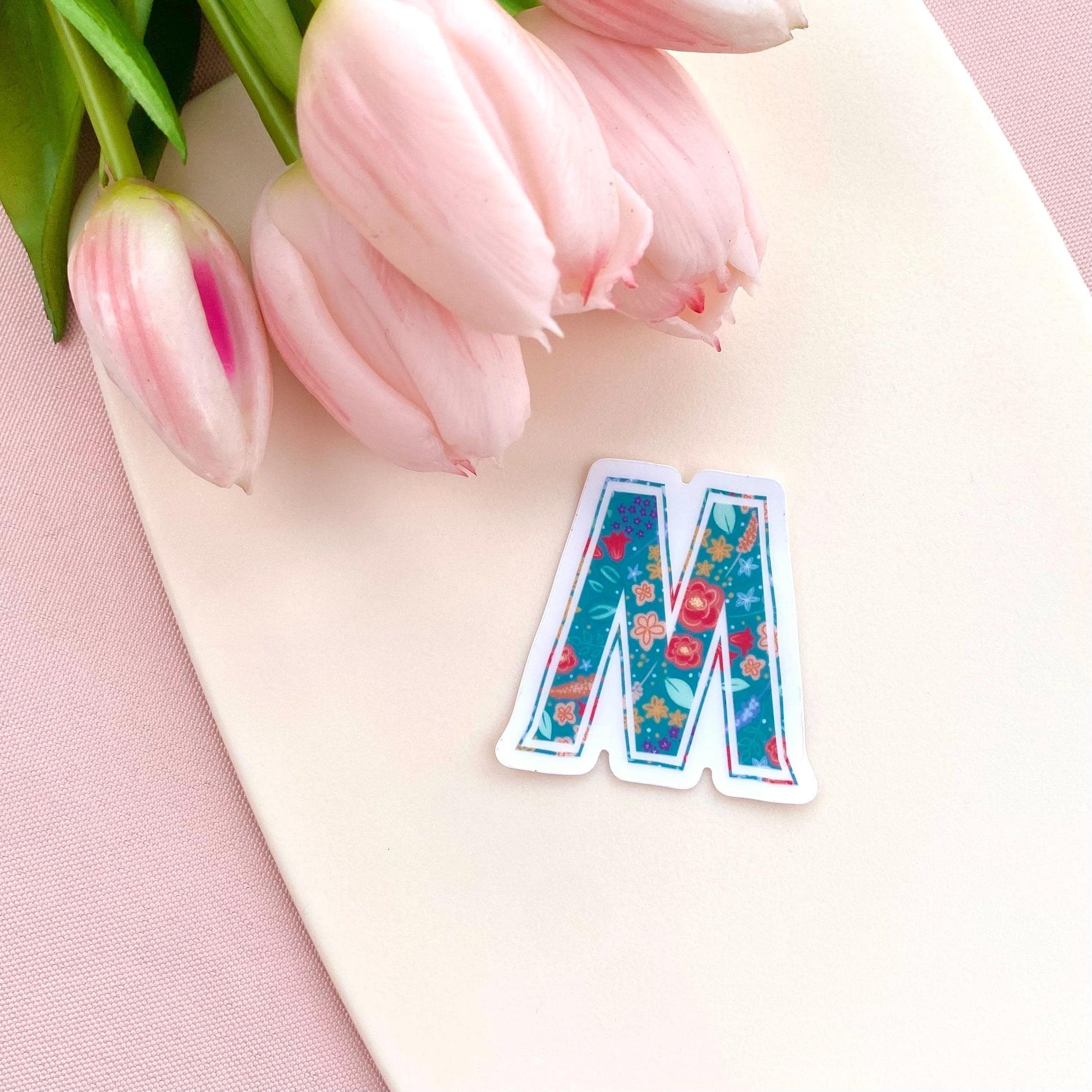Floral letter 'M' sticker on a pink surface with pink tulips