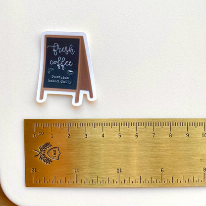 Sticker of a coffee shop sign with a chalkboard and a ruler for scale on a white background