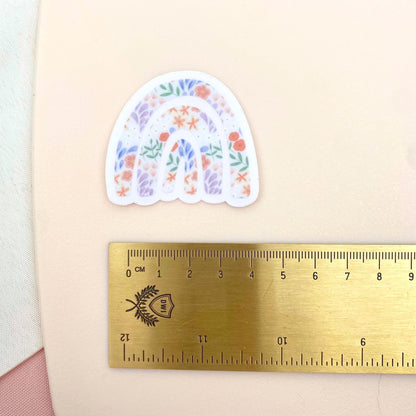 Decorative rainbow sticker with floral design next to a ruler on a beige background