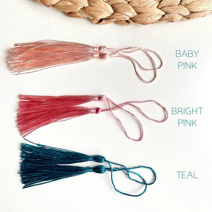 Three tassels in baby pink, bright pink, and teal on a white background with a woven mat.