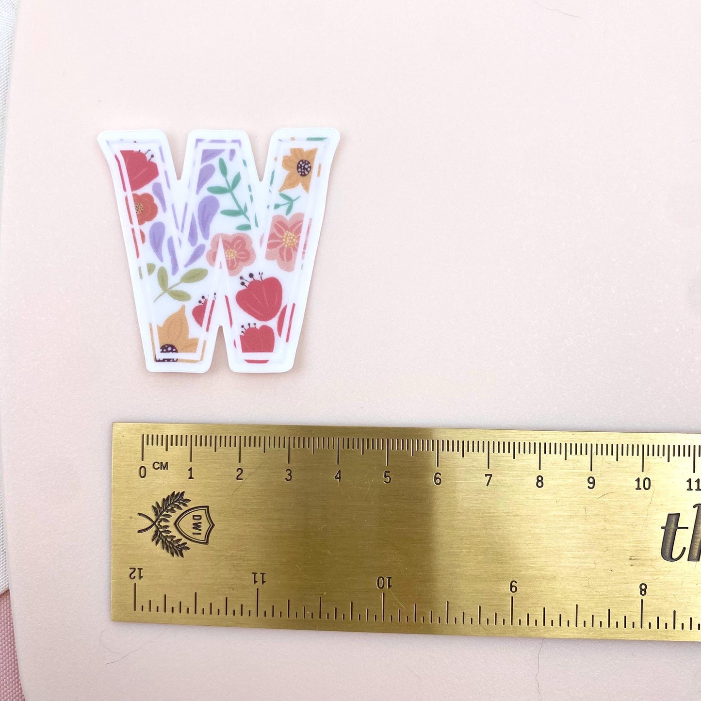 Sticker with floral design on a letter 'W' next to a ruler for scale on a pink background