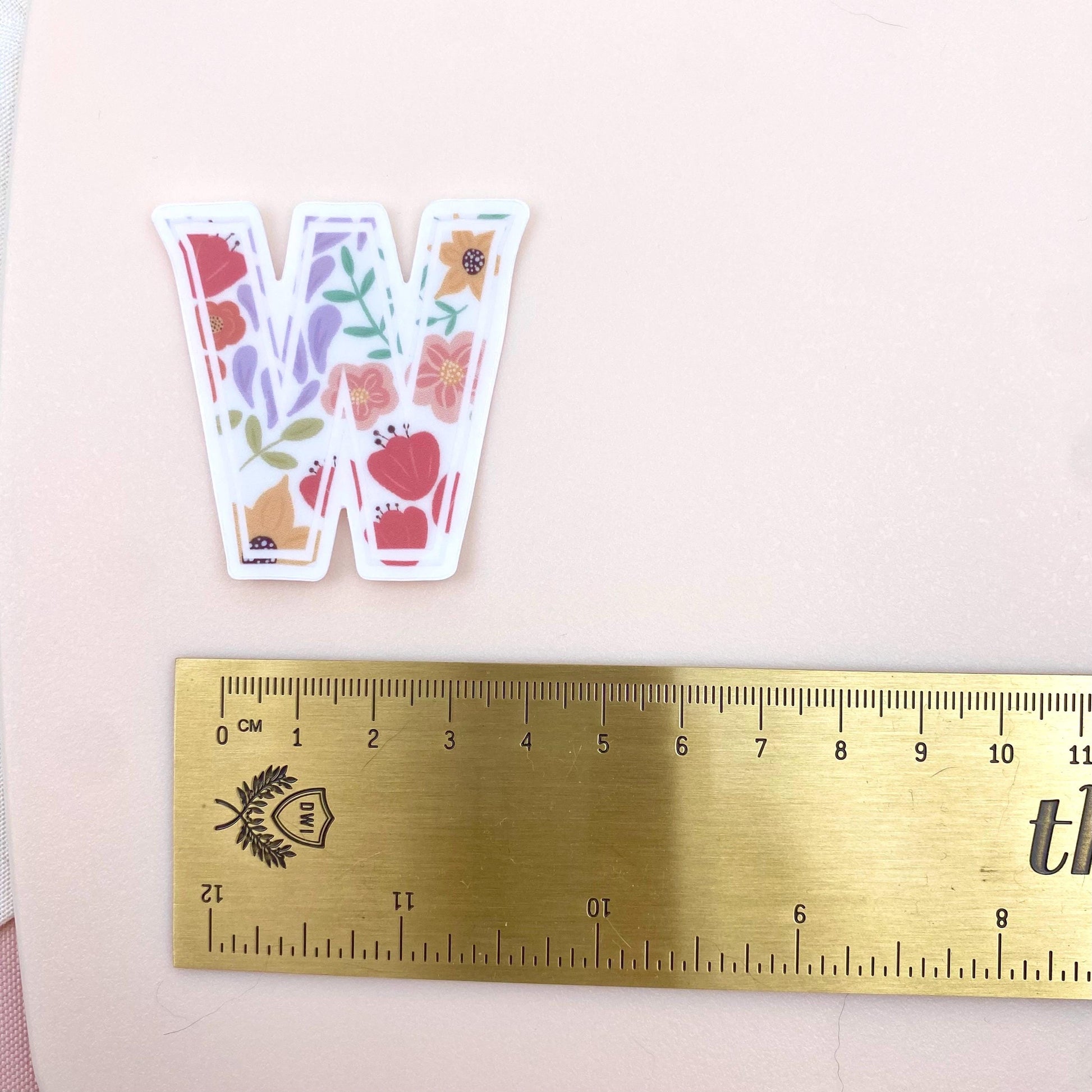 Sticker with floral design on a letter 'W' next to a ruler for scale on a pink background
