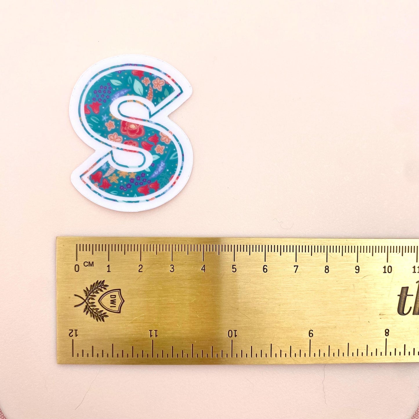 Floral letter 'S' sticker next to a ruler on a beige background