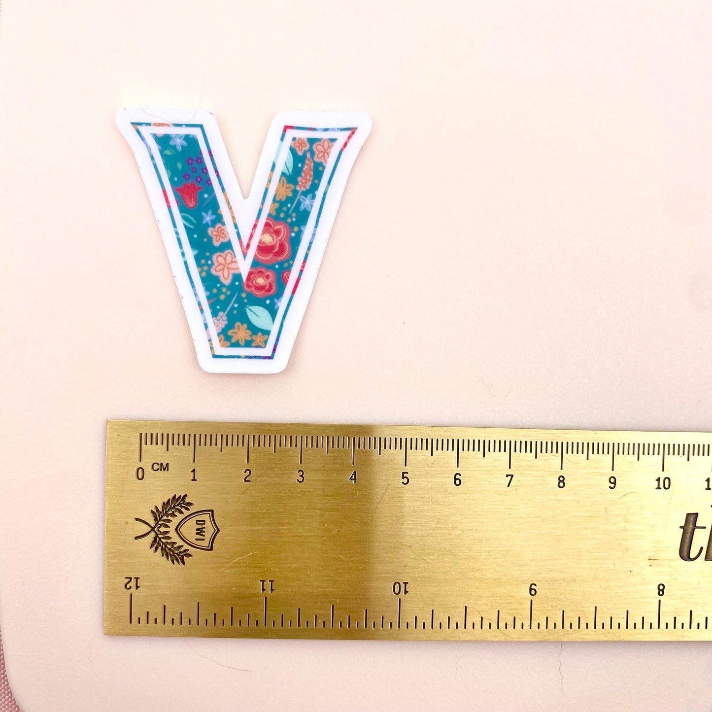 Floral letter 'V' sticker next to a ruler on a beige background