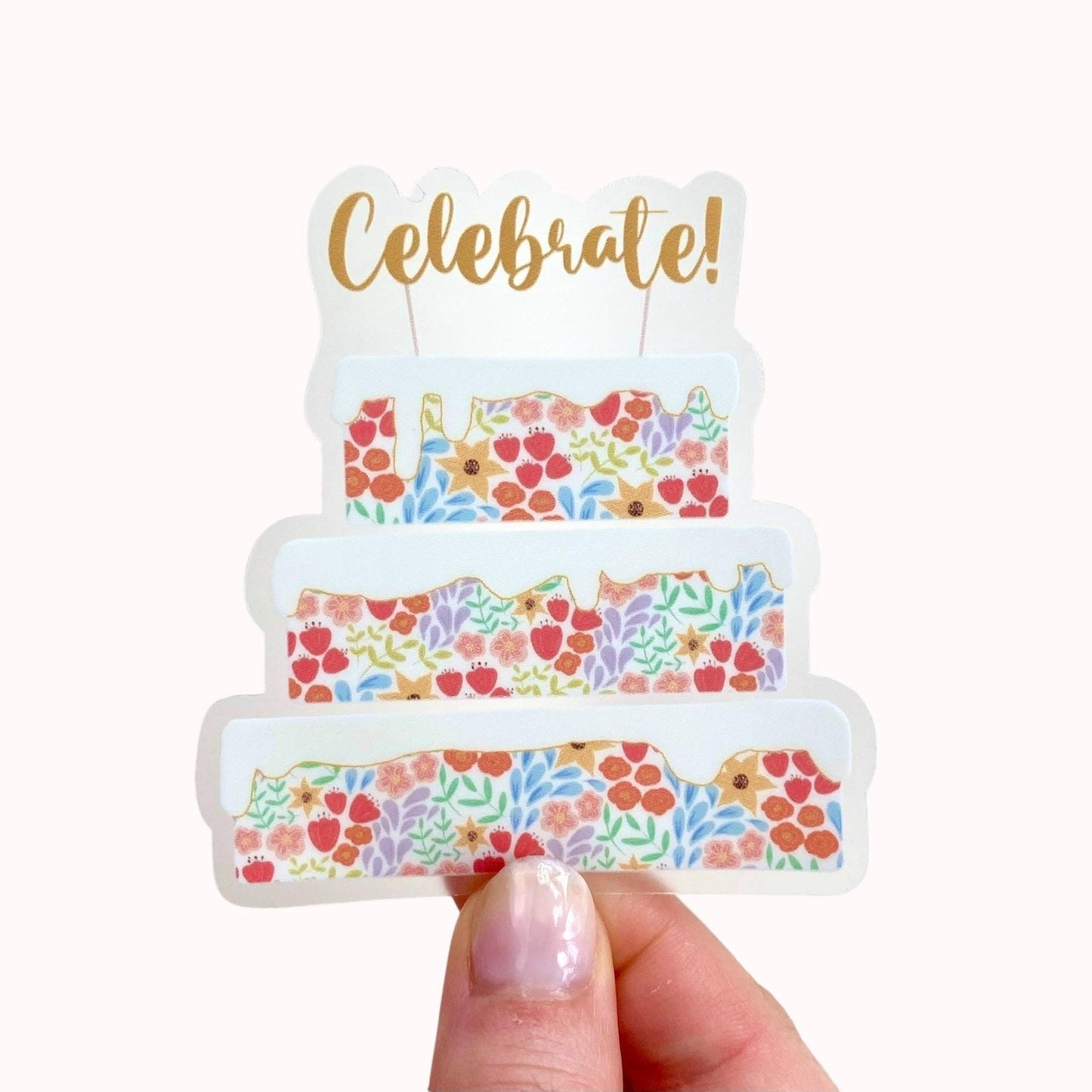 Decorative 3 tiered cake sticker with floral patterns and 'Celebrate!' text held by a hand on a white background