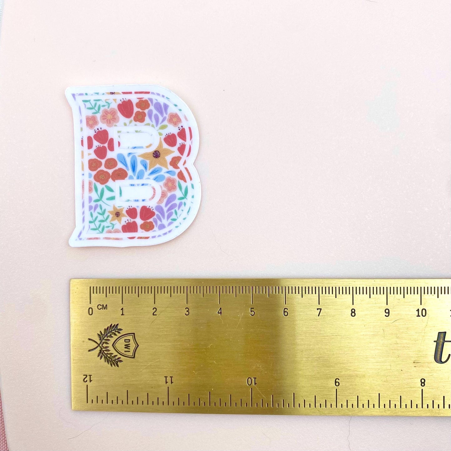 Floral letter 'B' sticker next to a ruler on a light pink background