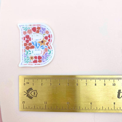 Floral letter 'B' sticker next to a ruler on a light pink background