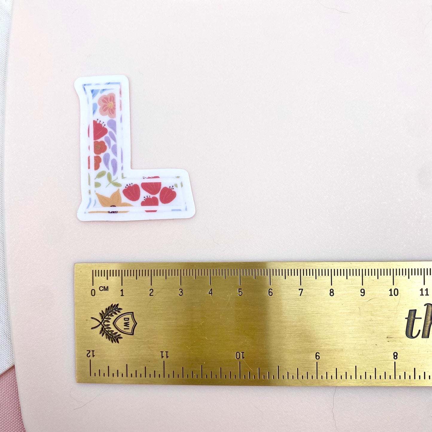 Floral letter 'L' sticker next to a ruler on a pink background